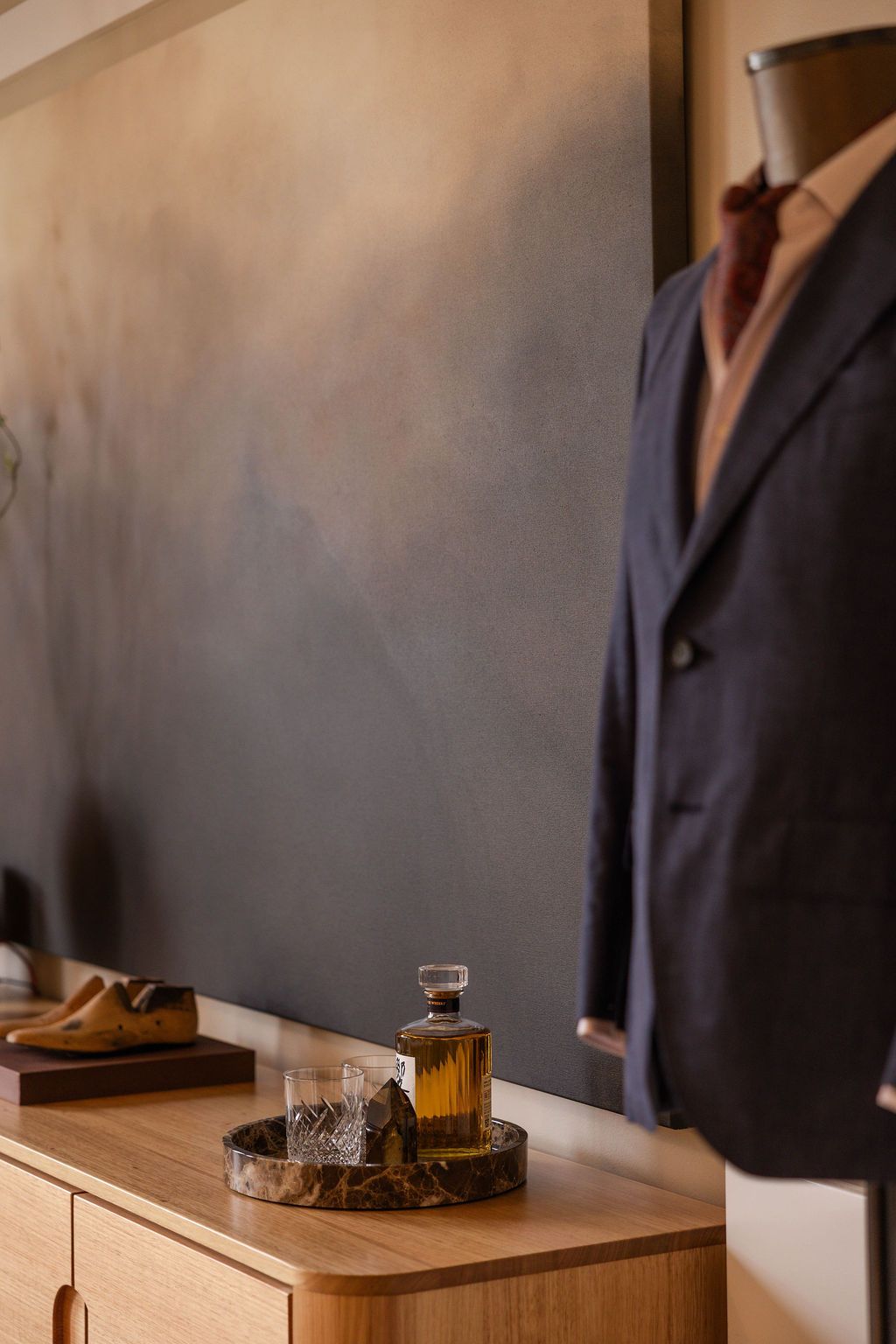 An out-of-focus suited mannequin in the foreground, in-focus is a whisky bottle alongside glasses atop a marbel platter, in front of a painting
